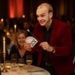 Ein Magier in einem roten Jackett zeigt Karten während einer Dinner-Show mit Gästen im Hintergrund.
